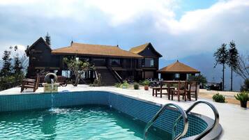 A pool area with a clear blue pool, wooden deck, and a large building with a thatched roof.