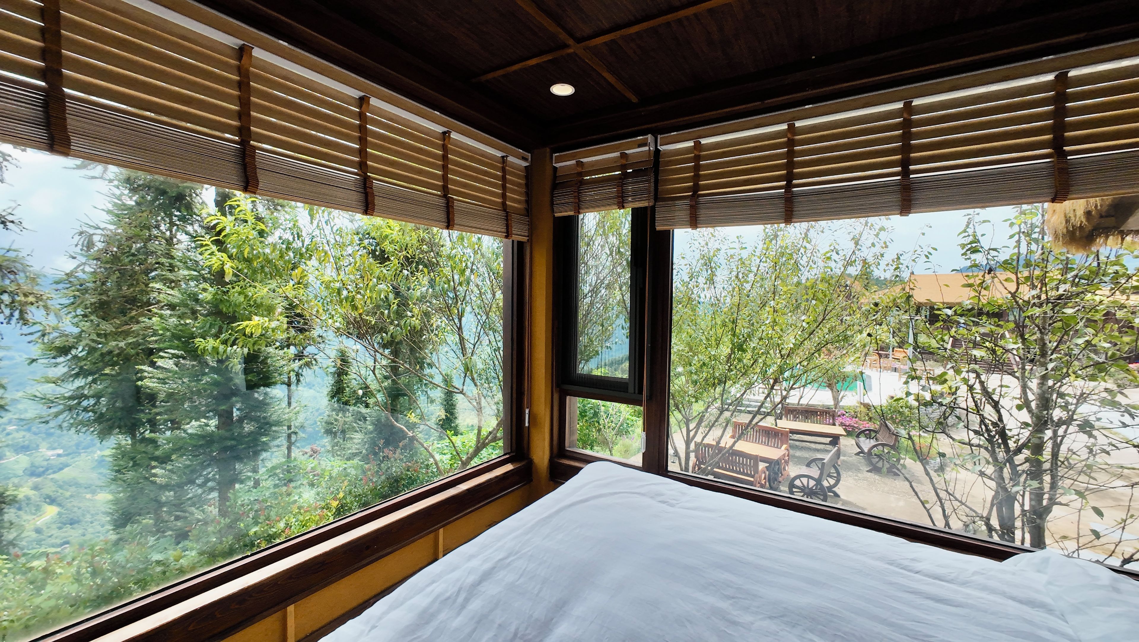 Quarto com grande janela, cama e vista para árvores e um prédio lá fora.