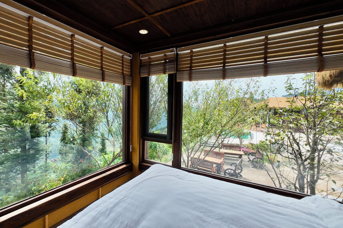 A room with a large window, a bed, and a view of trees and a building outside.
