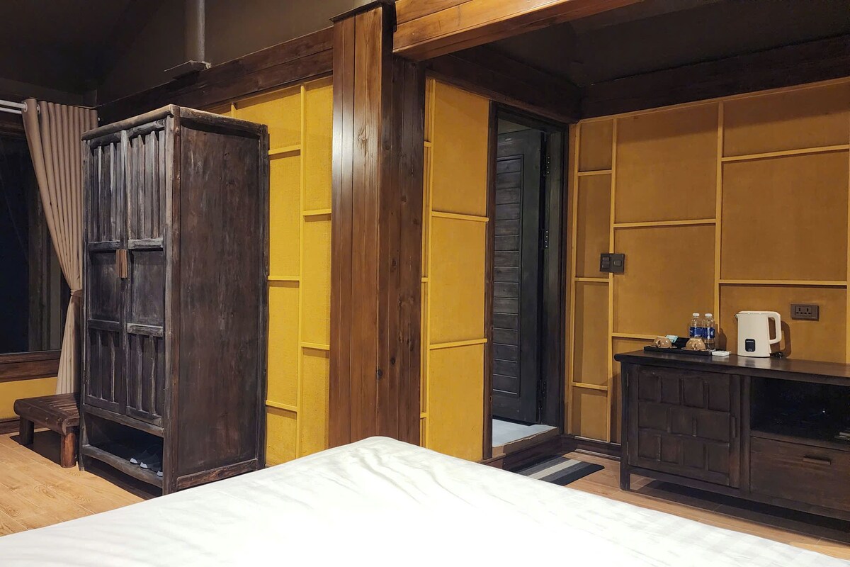 A hotel room with a bed, a wooden wardrobe, a bench, a small table with a kettle, and a window with curtains.