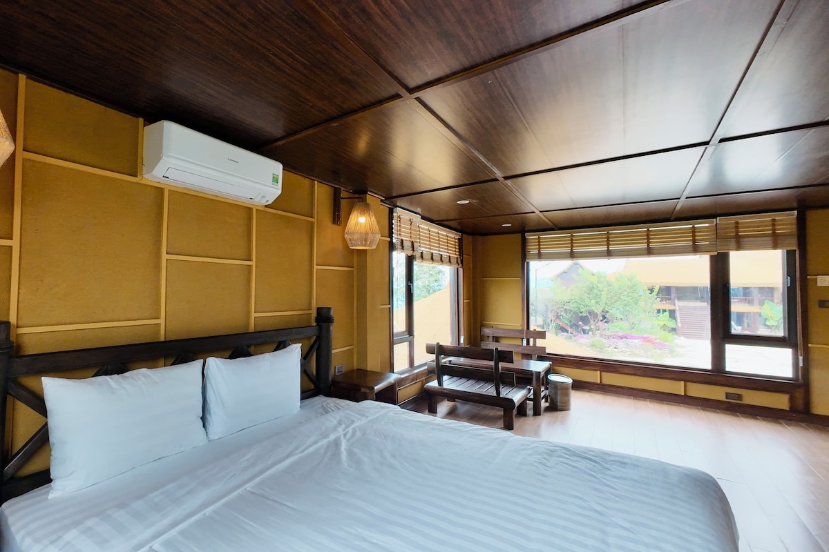Una habitación de hotel con una cama, cabecero de madera, una ventana con persianas, una mesita y una lámpara colgante.