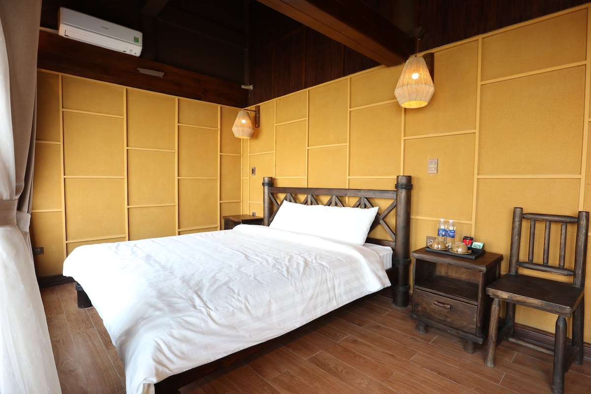 Habitación de hotel con cama, cabecera de madera, dos mesitas de noche, una silla y aire acondicionado de pared.