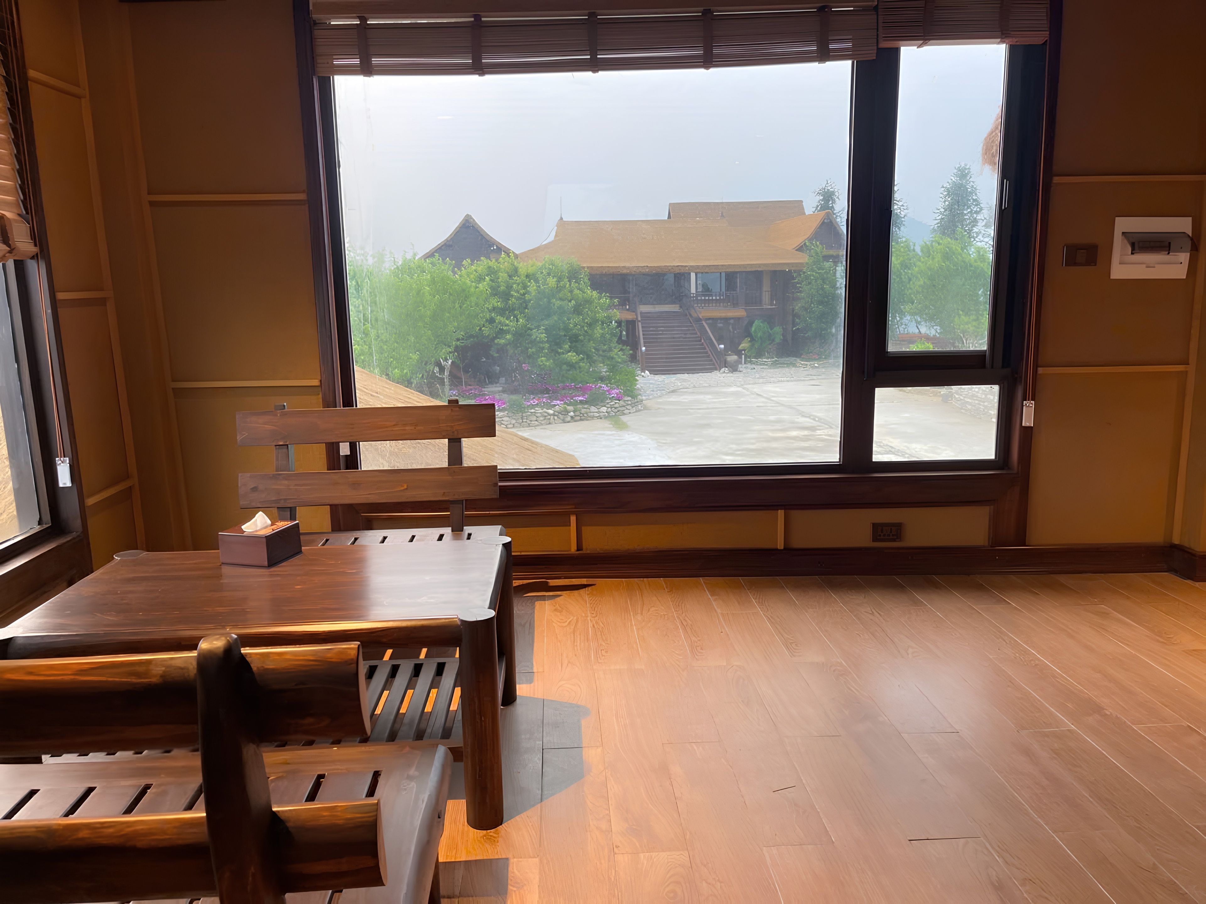 A room with a large window, wooden furniture, and a view of a building with a staircase.