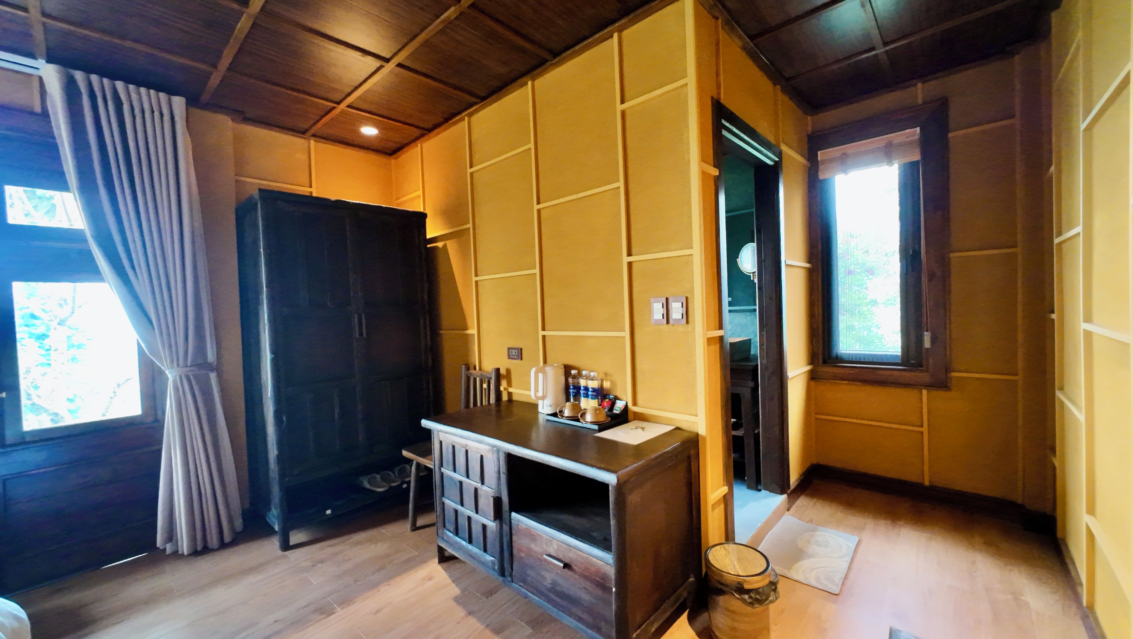A room with a wooden floor, a desk with a kettle and cups, a wardrobe, and a window with curtains.