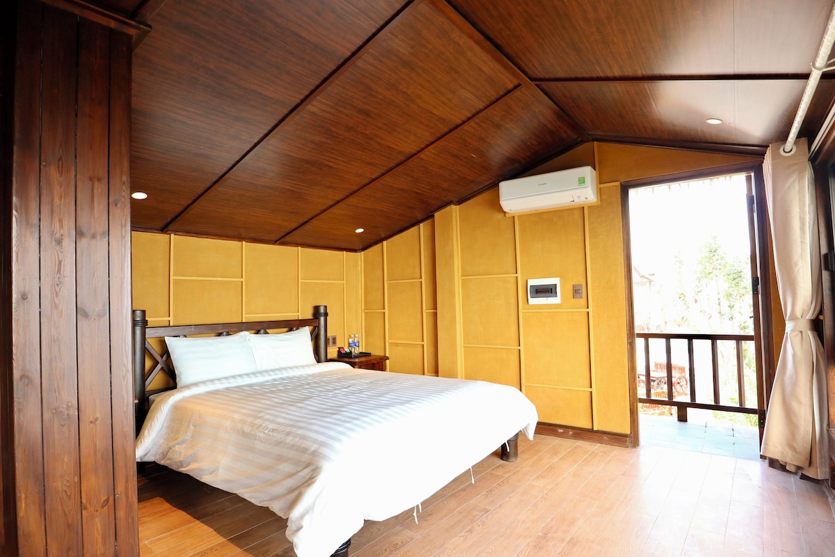 Habitación de hotel con techo de madera, cama con ropa blanca, mesita de noche, equipo de aire acondicionado y vista al exterior a través de una ventana.