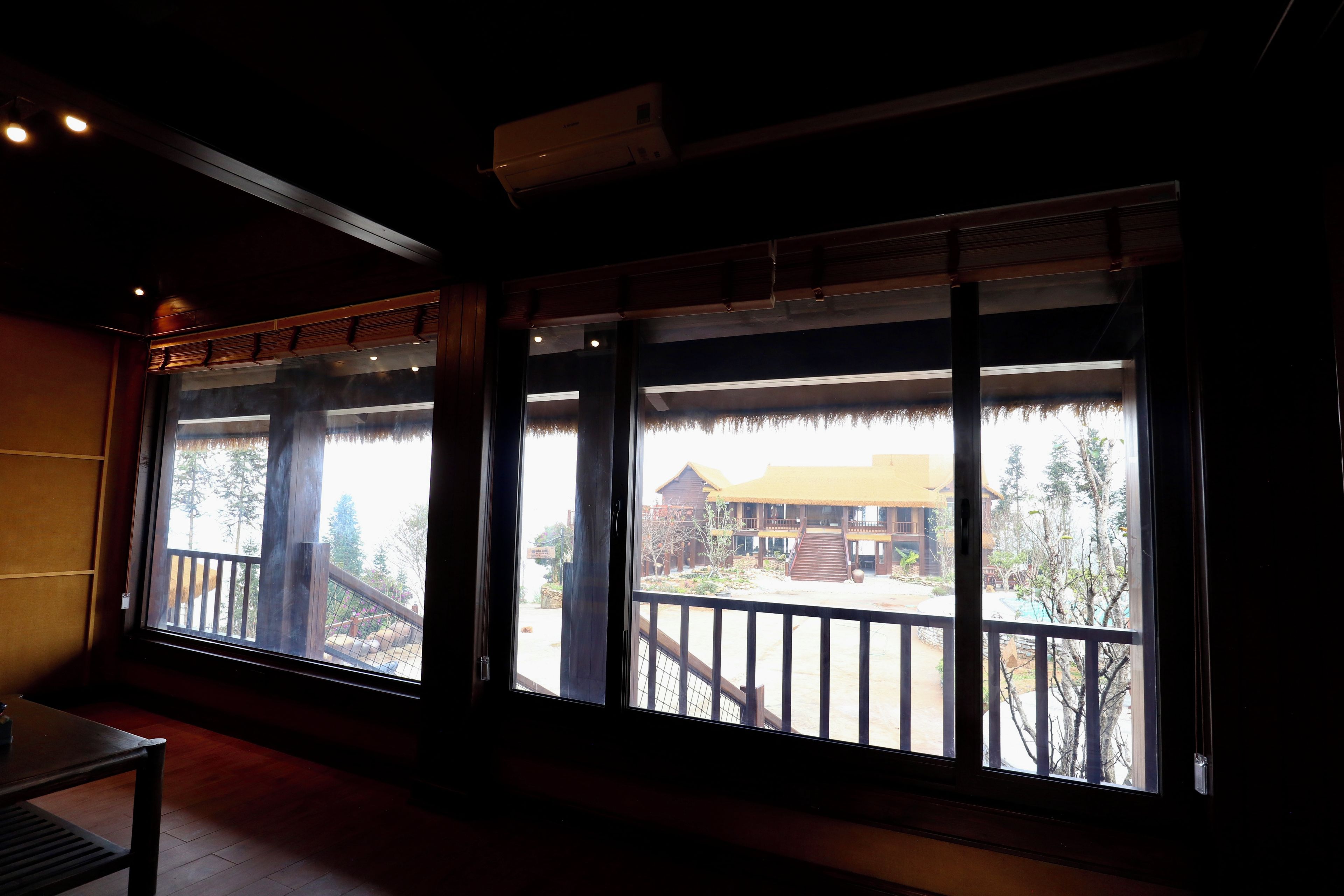 Una habitación con grandes ventanales que dan a un edificio con tejado de paja y una terraza de madera.