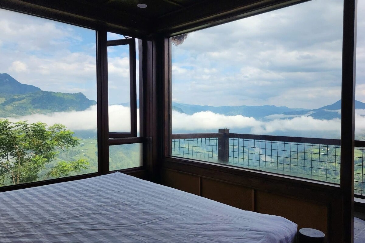 A room with a bed and a large window overlooking a mountainous landscape with clouds.