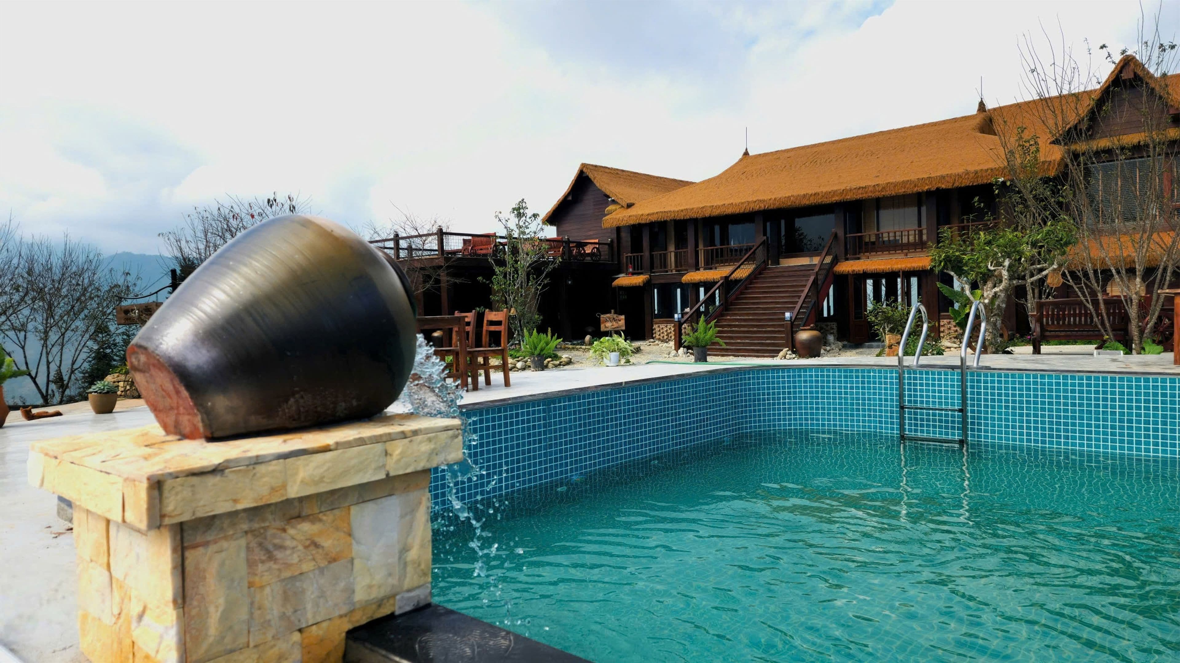 Una piscina con una estructura de piedra en un lado, una terraza de madera con sillas y un edificio con techo de paja al fondo.