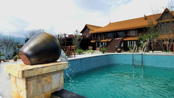 Una piscina con una estructura de piedra en un lado, una terraza de madera con sillas y un edificio con techo de paja al fondo.