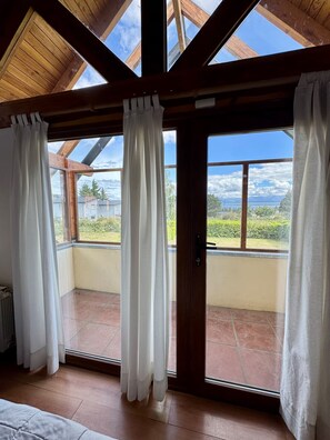 Quarto com teto inclinado, vigas de madeira e vista para uma paisagem verde através de amplas janelas com cortinas brancas.