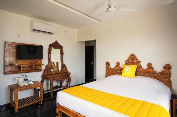 Una habitación de hotel con una cama, cabecera de madera, televisor montado en la pared, espejo y una mesita de noche con lámpara.