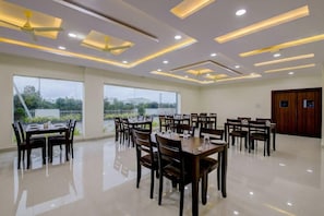 Un comedor amplio con varias mesas y sillas, ventiladores de techo, iluminación empotrada y ventanas grandes que ofrecen vistas a zonas verdes y edificios.