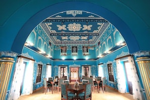Une pièce dotée d’un plafond bleu et blanc richement décoré, d’une table centrale entourée de chaises et offrant une vue sur l’extérieur par le biais d’une fenêtre.