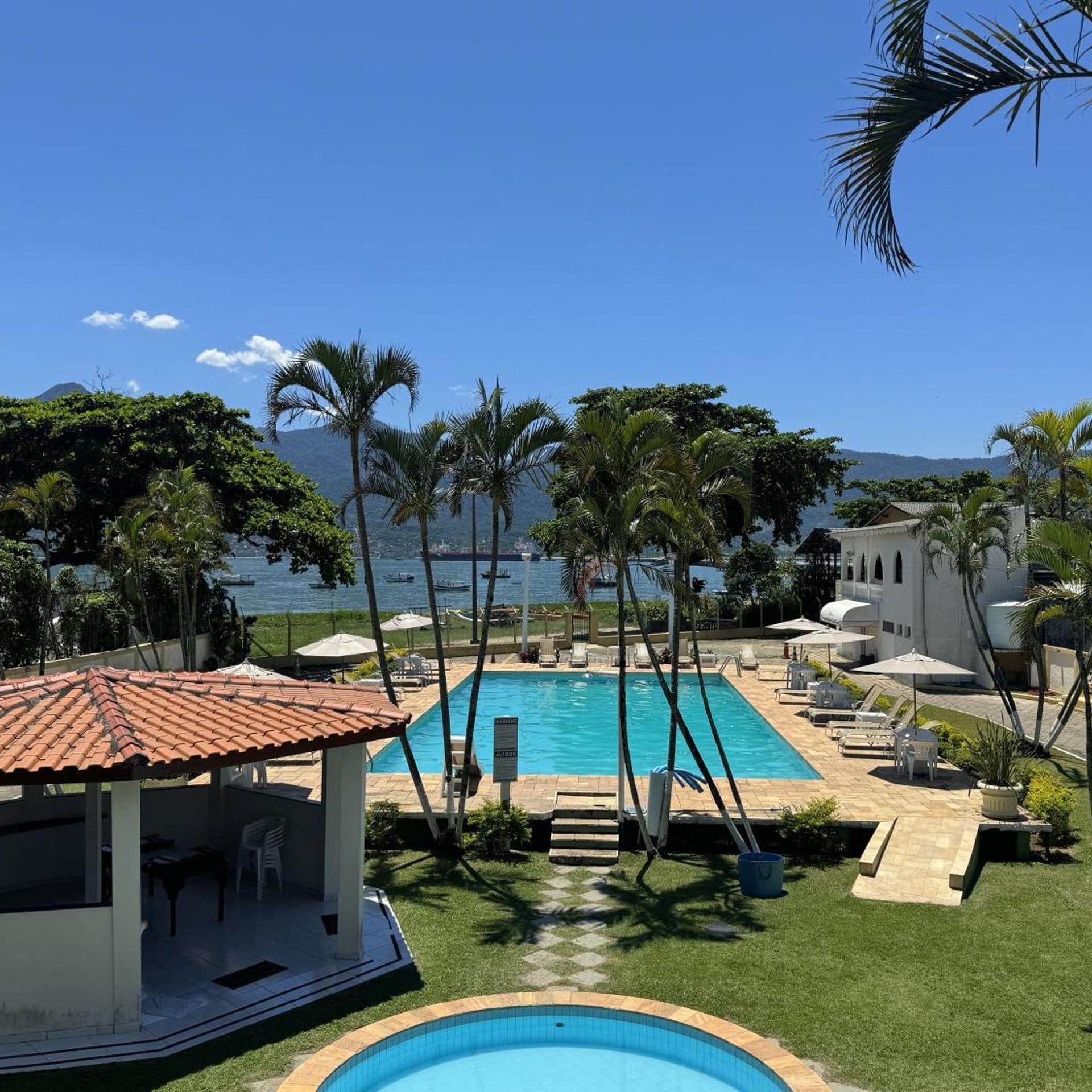 A pool area with a view of the sea, surrounded by palm trees and a building with a tiled roof.