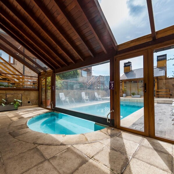 Una piscina interior con suelo de piedra, vigas de madera en el techo y puertas de cristal que dan a un área exterior con sillones de descanso.
