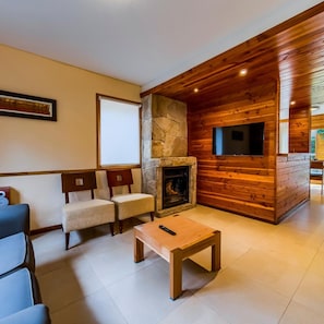 Una sala de estar acogedora con chimenea de piedra, paredes de madera, televisor de pantalla plana y un área de descanso con dos sillones y un sofá.
