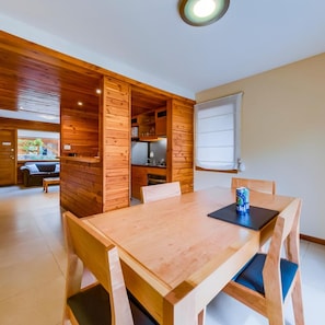 Una mesa de madera con sillas, una zona de cocina y un ventanal con persianas.