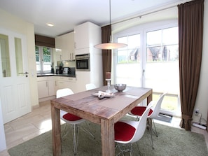 A dining area with a wooden table and red cushioned chairs, a kitchen with white cabinets, and a view through a glass door. - Theodor-storm-str. 3a (Sylt)