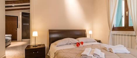 Een hotelkamer met een bed, nachtlampen, een nachtkastje, een raam met gordijnen en een deur naar een andere kamer.