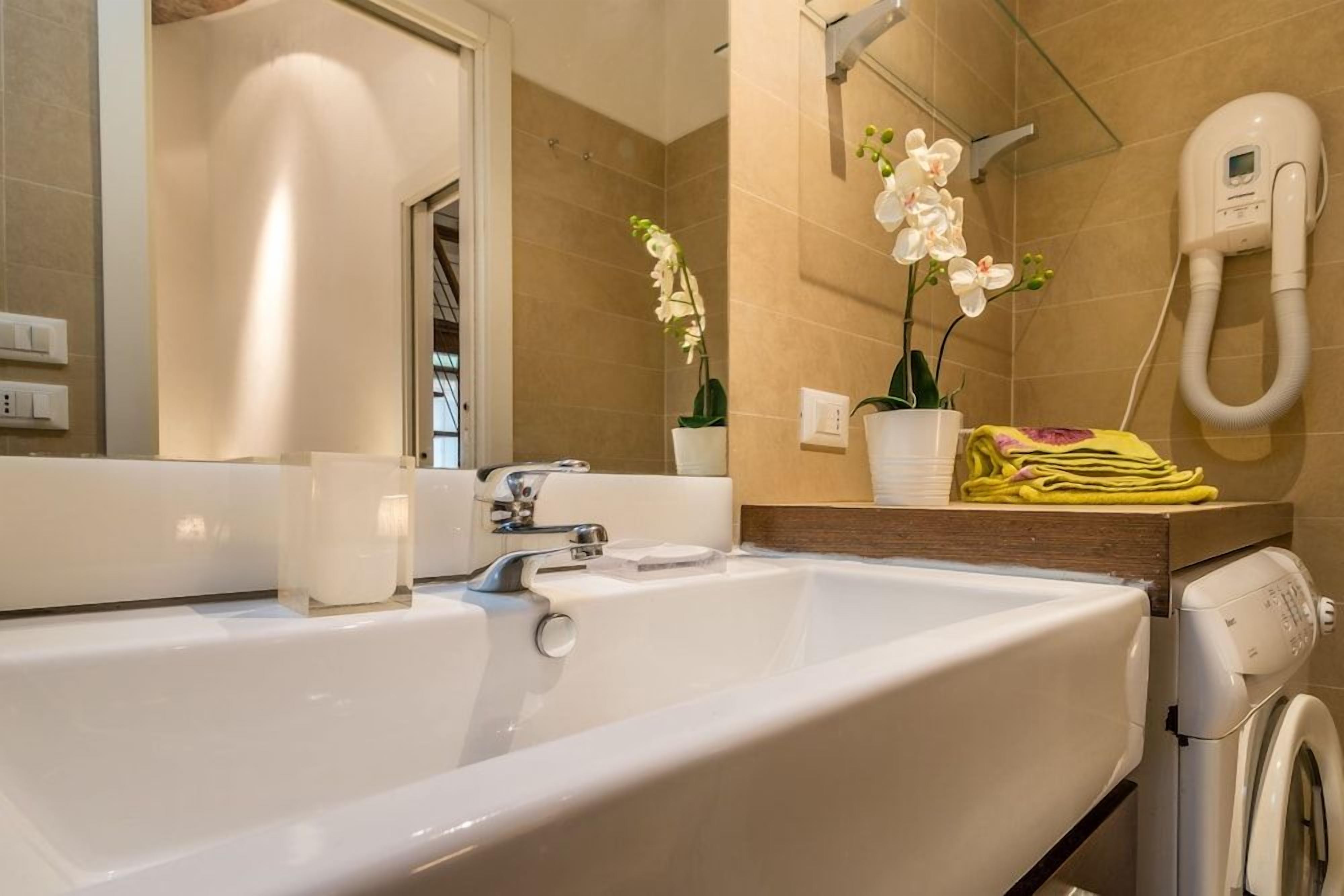 Un baño con una bañera blanca grande, un espejo, un lavabo blanco, un secador de pelo y una orquídea en maceta.