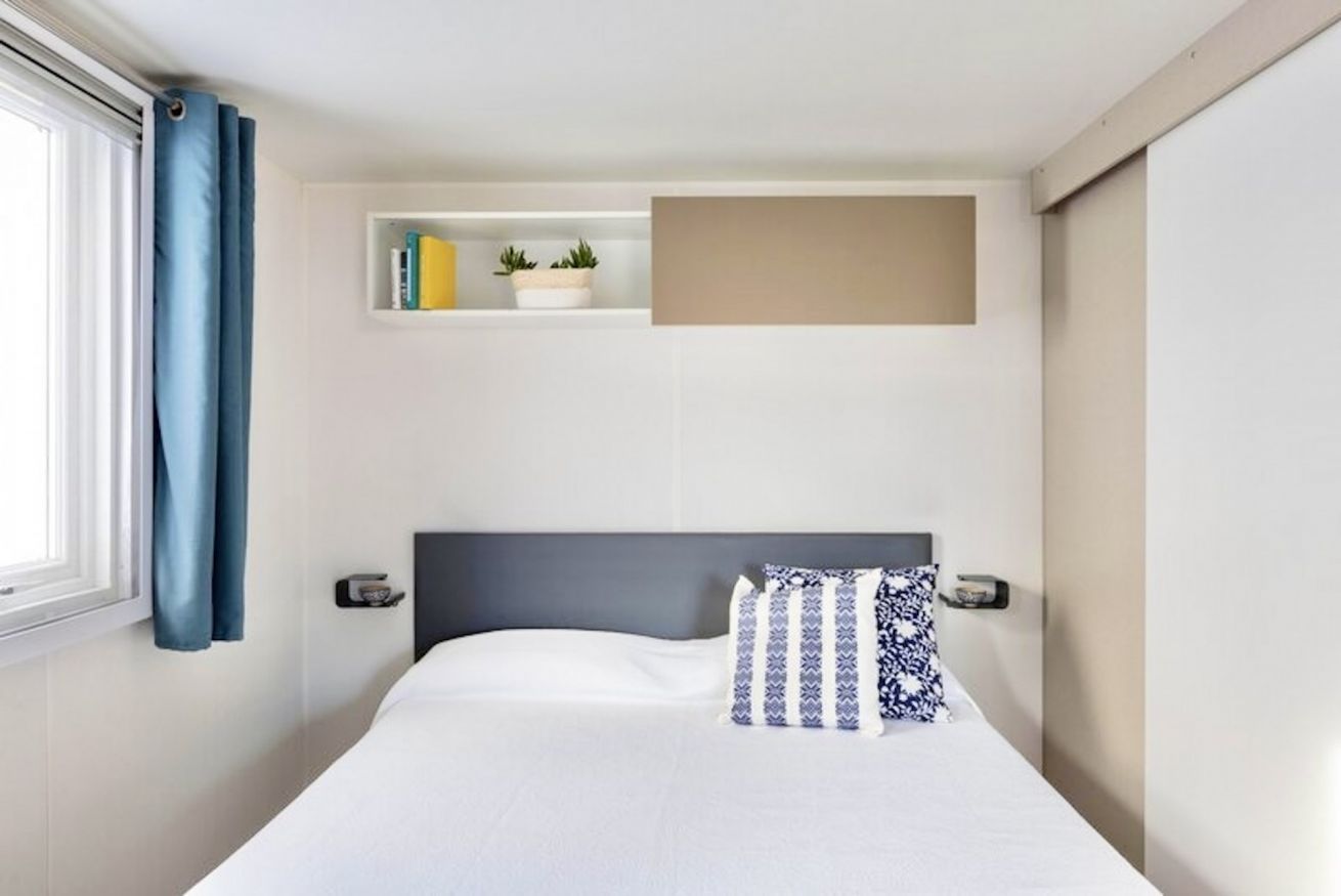 A hotel room with a bed, a window with curtains, a shelf with books and plants, and a wall-mounted panel.