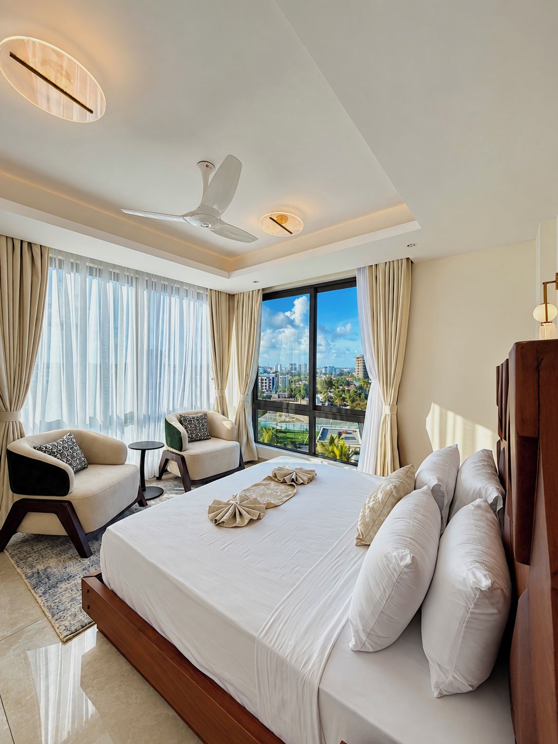 A hotel room with a large bed, a sitting area with a chair and a small table, a ceiling fan, and a view of the city through large windows.