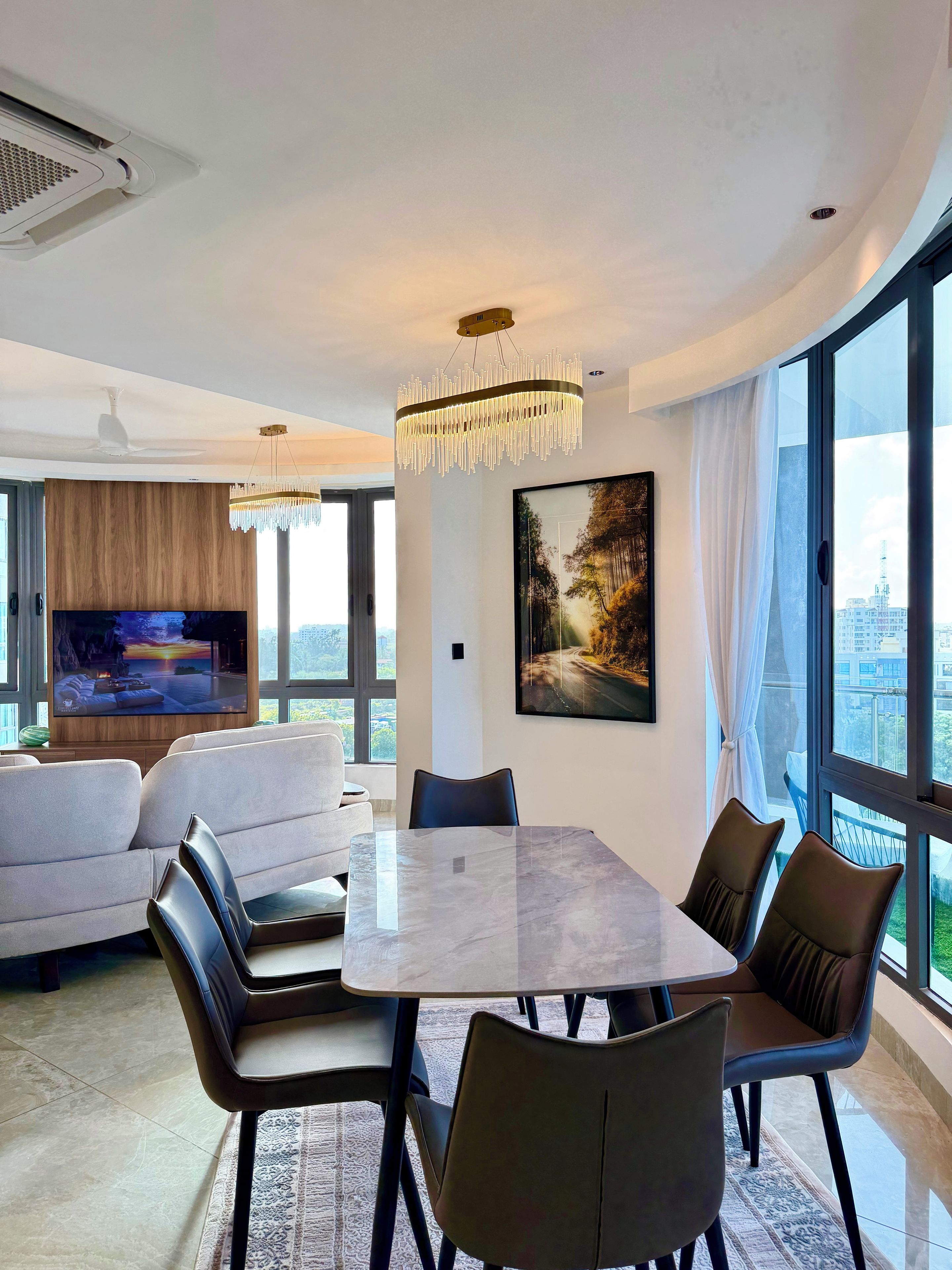 A modern living room with a large flat-screen TV, a dining area with a marble table, and a city view through large windows.