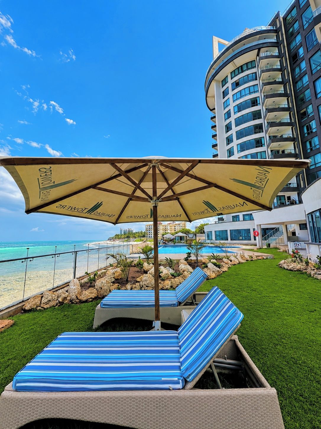 A poolside area with lounge chairs and an umbrella, a clear view of the ocean, and a modern building in the background.