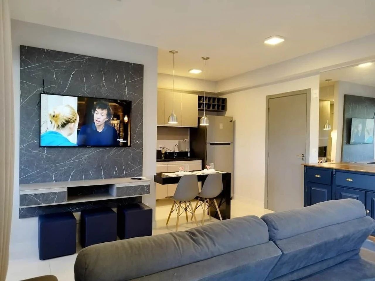 A modern living space with a flat-screen TV mounted on a marble wall, a grey sofa, a dining area with chairs, and a kitchen with cabinets and a refrigerator.