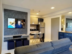 Un espacio de estar moderno con una televisión de pantalla plana montada en una pared de mármol, un sofá gris, un comedor con sillas y una cocina con armarios y un refrigerador.