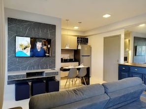 A modern living room with a flat-screen TV, a grey sofa, and a kitchen area with a dining table and chairs. - Unique Residence (São José do Rio Preto)
