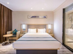 Habitación de hotel con una cama grande, mesitas de noche, una pequeña mesa redonda, un sofá y zona de ducha con mampara de vidrio.