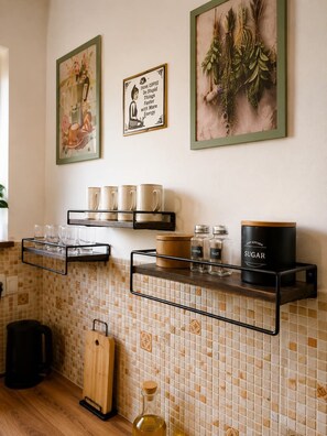 A kitchen with a tiled backsplash, a coffee station with mugs and a coffee machine, and a shelf with kitchen essentials like pepper and sugar.