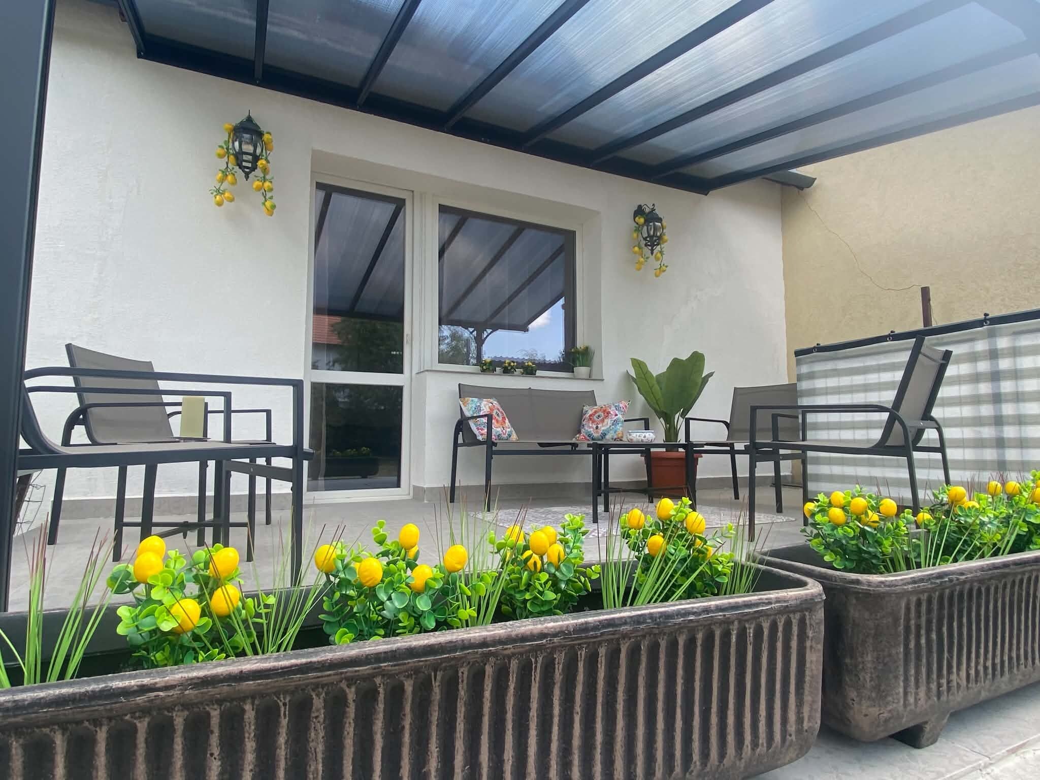 Un patio exterior con una zona de estar cubierta, plantas en macetas y una ventana con vistas.