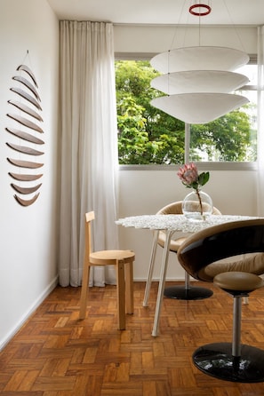 A modern dining area with a round table, two chairs, a hanging light fixture, and a window with a view of greenery. - Elegância no Jardim Paulista | 2 Quartos | 1 Vaga (São Paulo)