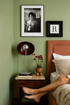 A bedroom with a bed, a bedside table with a lamp, a framed picture, and a framed letter on the wall. - Elegância no Jardim Paulista | 2 Quartos | 1 Vaga (São Paulo)