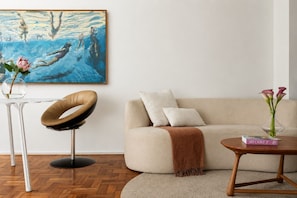 A modern living room with a beige sofa, a round coffee table, a unique chair, a painting on the wall, and a vase with flowers. - Elegância no Jardim Paulista | 2 Quartos | 1 Vaga (São Paulo)