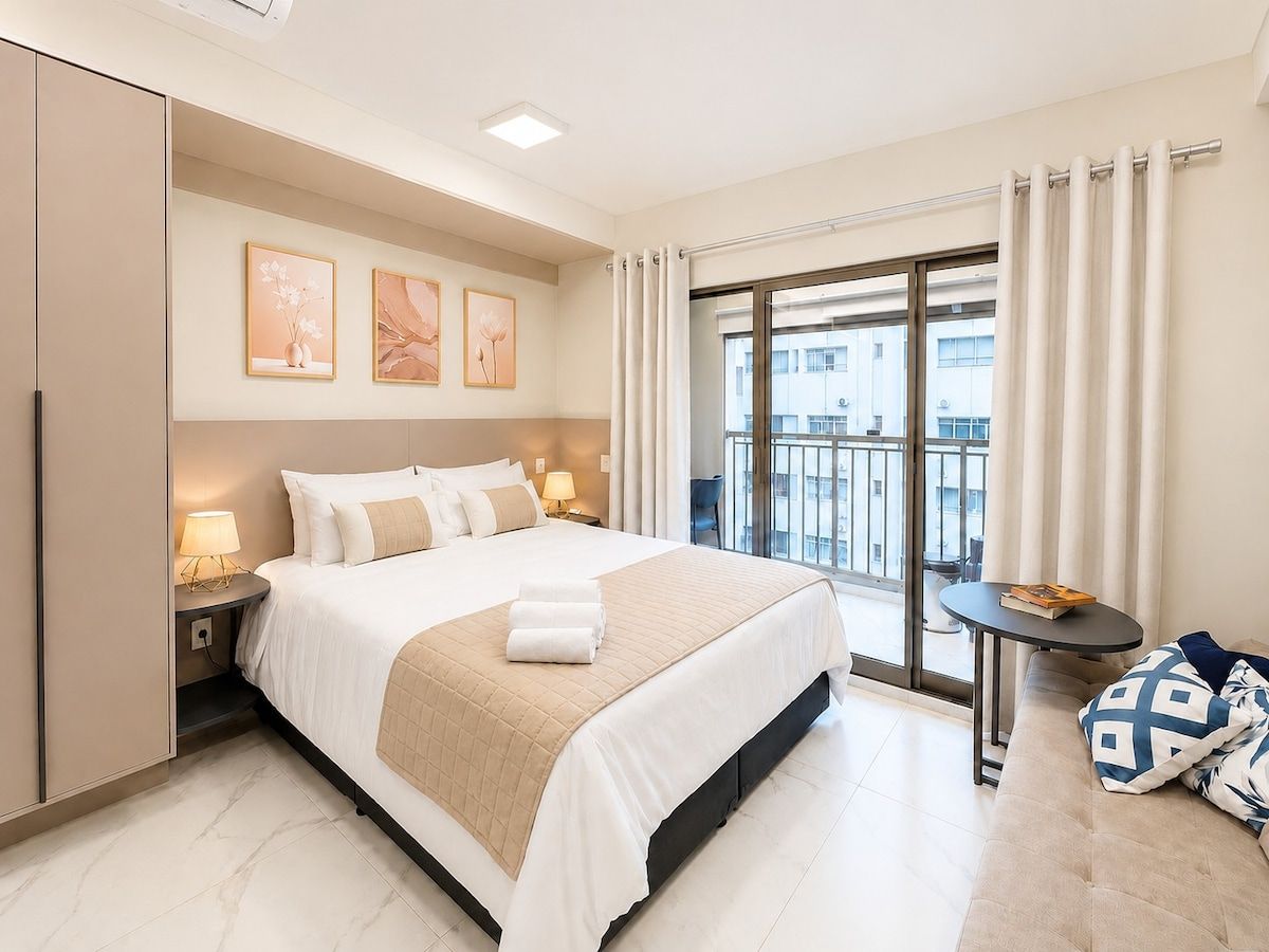 A hotel room with a large bed, a balcony with curtains, a small table, and artwork on the wall.