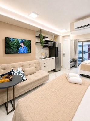 Ein modernes Hotelzimmer mit einer Couch, einem Bett, einem kleinen Tisch, einem Fernseher, einer Küchenzeile und einem Balkon.