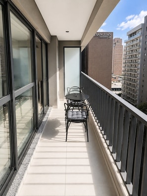 A balcony with a table and chairs, a glass door, and a view of a cityscape. - Conforto para até 4 pessoas | Metrô República (Santa Ifigênia)