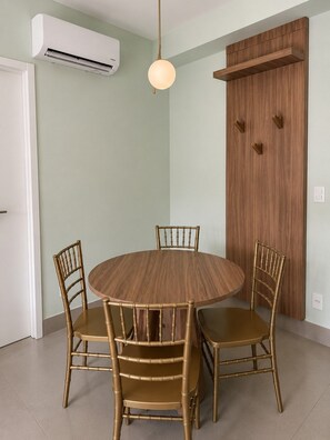 Uma área de refeições com uma mesa redonda e quatro cadeiras, um aparelho de ar condicionado, uma porta de madeira e um lustre.