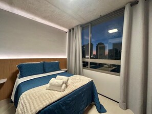 Een hotelkamer met een bed, nachtkastje, raam met gordijnen en uitzicht op de stad.