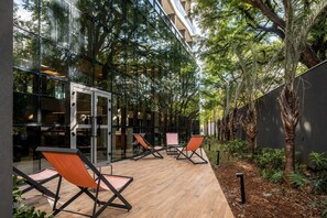 An outdoor seating area with chairs and a wooden deck, surrounded by glass walls and trees. - Av. Paulista | Metrô Paraíso | Conforto e Lazer (São Paulo)
