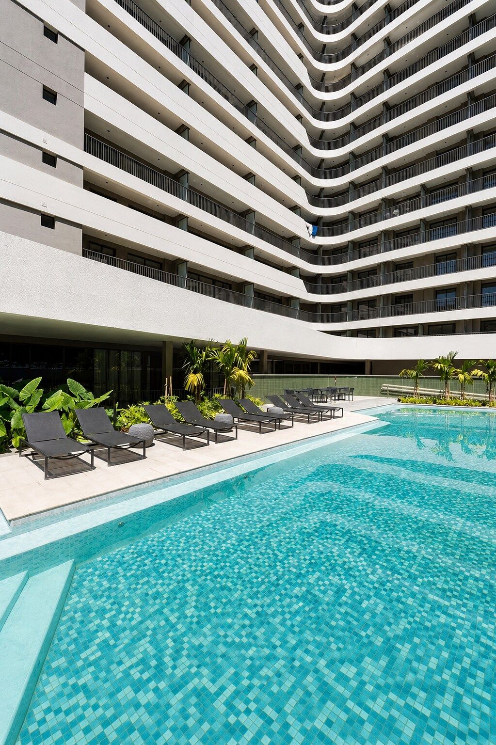 Un hotel moderno con zona de piscina de varios niveles, sillas de descanso y piscina de color azul cristalino.