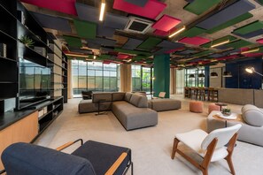 A modern lounge area with a sectional sofa, a flat-screen TV, and a bar area. - Studio Aconchegante | 16º Andar | Metrô República (Santa Ifigênia)