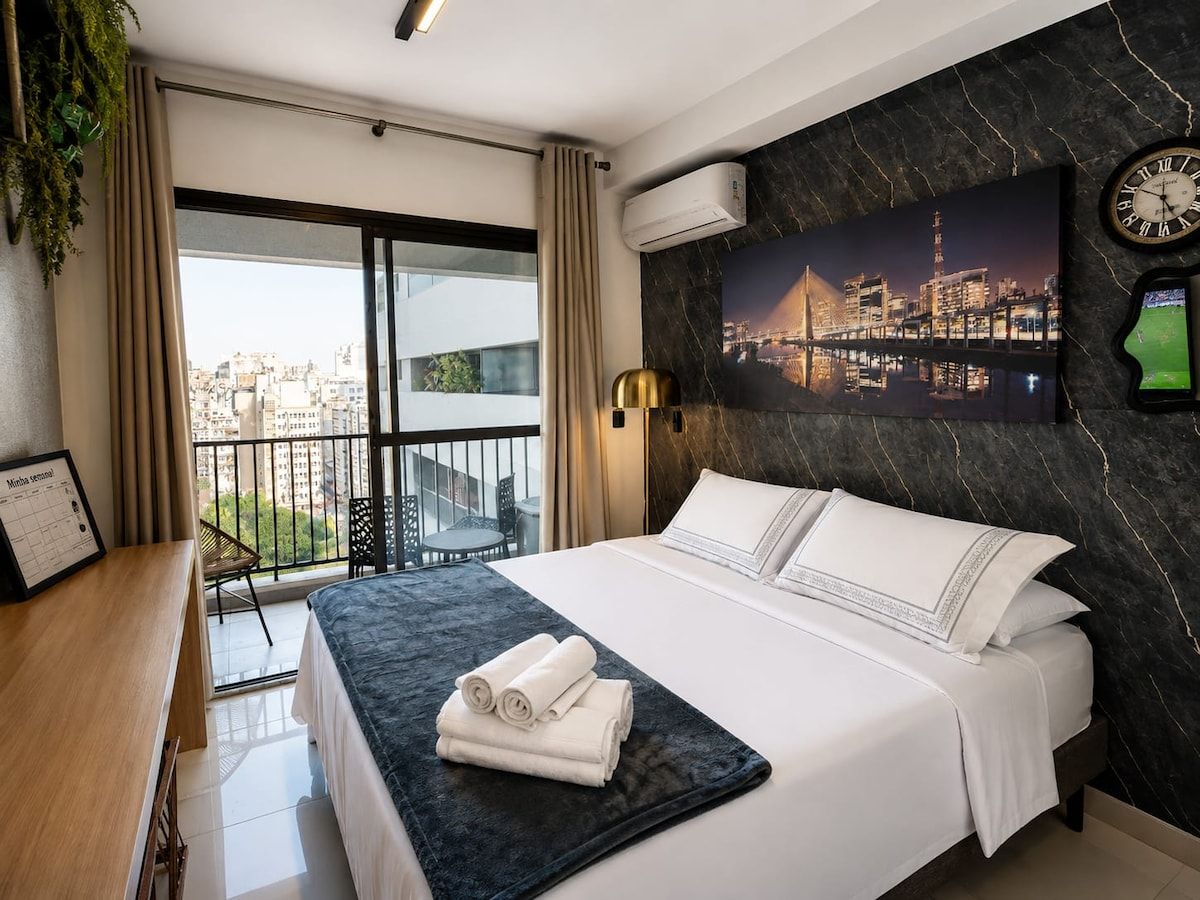 A hotel room with a bed, a desk, a chair, a balcony with a view of the city, and an air conditioning unit.