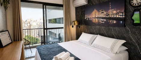A hotel room with a bed, a desk, a chair, a balcony with a view of the city, and an air conditioning unit.