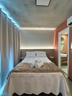 Une chambre d’hôtel avec un lit, des rideaux, une table de chevet, un miroir et une salle de bain visible à travers une porte ouverte.