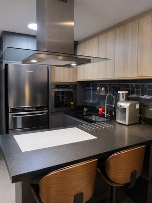 A modern kitchen with stainless steel appliances, a black countertop, wooden cabinets, and a built-in island with bar stools. - Moderno e Equipado | 1 Vaga | Metrô República (São Paulo)