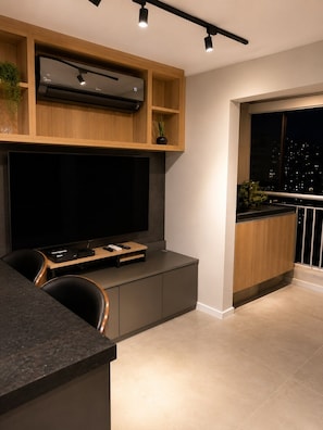 Ein modernes Wohnzimmer mit großem Flachbildfernseher, integrierten Regalen, einer Küchenzeile und einem Balkon mit Stadtblick.